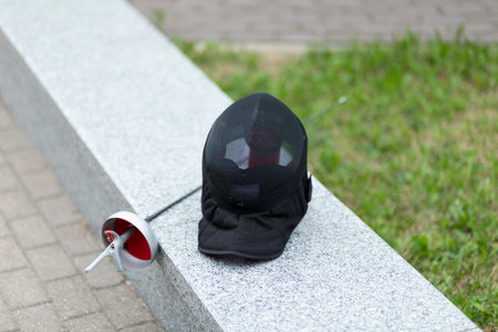 Metal epee and black fencing helmet are laying on border outside in the park