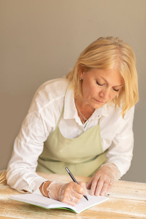 White beautiful caucasian blonde mature woman makes notes in notebook (planner) and on wooden table,...