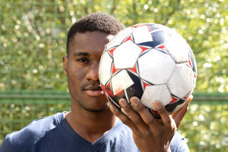 Young confident african american guy hides an eye by soccer ball outside, on stadium. Focus on...