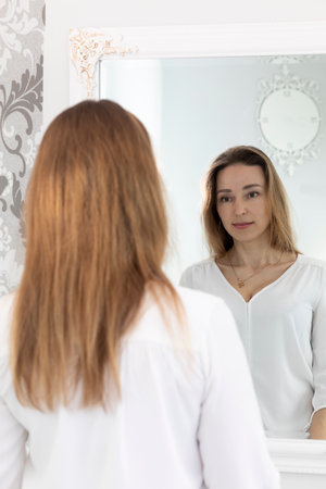 Portrait Of Charming Beautiful Brunette Caucasian 40 Yo Woman Looking At Mirror And Smiling. Healthy Moisturized Skin Care, Aging Beauty, Skincare Treatment Cosmetics Concept. Vertical Plane.の写真素材