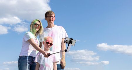 Caucasian Family Takes Selfie. Parent And Child With Colorful Dye At Birthday Party Or Celebrating Holi Color Festival, Blue Sky On background. Cheerful Family Spend Time Together. Horizontal Planeの写真素材