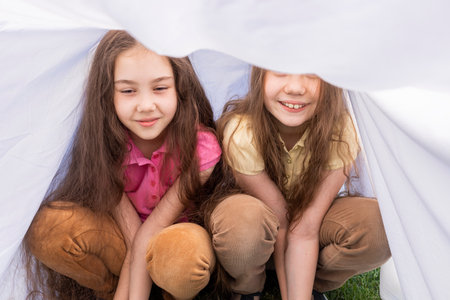 Two Little Girls, Sisters With Long Hair Sits Under Sheet On Grass in Meadow. Caucasian Asian Siblings Having Fun, Joy Together. Family Love And Care, True Friendship. Horizontal Plane.の写真素材