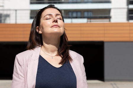 Relaxed Mature Woman With Closed Eyes Enjoys Sun, Peace In Apartment Campus, Complex In Daytime. Female Brunette Wears Casual Clot. Modern Residential Building, Horizontal Planeの写真素材