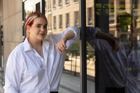 Portrait Young Real Female Smiling And Standing Near Glass Window Of Modern Building In City Looking At Camera. Carefree Beautiful Plus Size Adult, Student. Copy Space. Horizontal Planeの写真素材
