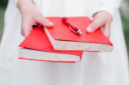 a girl in white holds books in a red cover in her hands on books lies a red penの写真素材