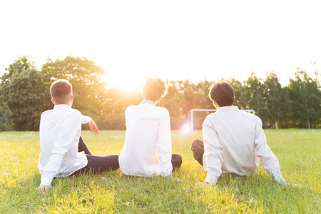 Three boys are sitting with their backs to us on the soccer field in white shirts at sunsetの写真素材
