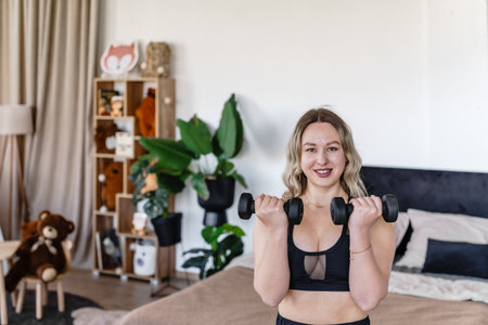 Female trainer in sports top with dumbbells in hands training online at home in bedroomの写真素材