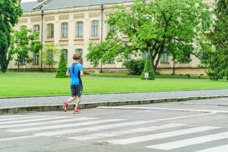 A male athlete runs in the park near buildings along the road in summerの写真素材