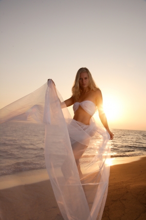 Beautiful young woman on the beach wrapped in white wedding veil at sunriseの写真素材