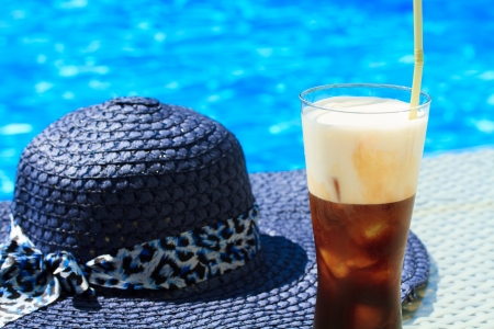 Ice coffee Fredo against blue clear water of the swimming pool with straw hatの写真素材