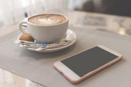 In the cafe, a Cup of delicious coffee with a heart of coffee foam, next to a white phone with a black screen. Remote work, work from homeの写真素材