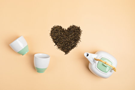 White ceramic tea pot with dry green tea leaves in heart shape on beige background. Eco friendly organic brand concept. Branding mockupの写真素材