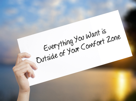 Everything You Want is Outside of Your Comfort Zone Sign on white paper. Man Hand Holding Paper with text. Isolated on sunset background.  technology, internet concept.の写真素材