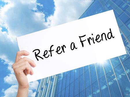 Refer a Friend  Sign on white paper. Man Hand Holding Paper with text. Isolated on Skyscraper background.  Business concept. Stock Photoの写真素材