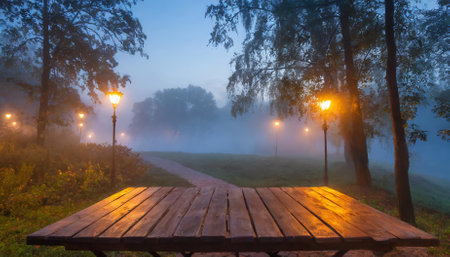 A picturesque wooden table stands in a quiet park enveloped by gentle evening fog. The misty atmosphere, softened by dim light, creates a peaceful scene filled with rustic charm and naturalの素材