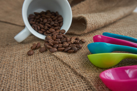 Loose roasted coffee beans, white porcelain coffee mug and color plastic dose measuring spoons on jute sack. Conceptual of recommended amount of coffee per day. Low angle selective focus and copy spaceの写真素材