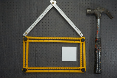 Conceptual construction and renovation still life with a flat lay design of a house formed by a yellow and white rulers, hammer and blank sticky notes on a black perforated backgroundの写真素材