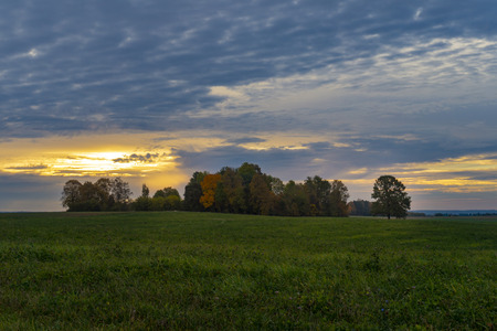 Cloudy sunset over green field and autumn colored treesの写真素材