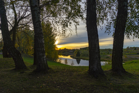 Golden sunburst through the green leaves of a birch trees on the shores of a tranquil river surrounded by woodland in a scenic landscapeの写真素材