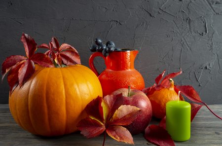 Two pumpkins with bright autumn leaves apple, grapes red jug and green candle on wooden rustic table against a black rough concrete grunge wall. Autumn Harvest and Holiday still life with free copy spaceの写真素材