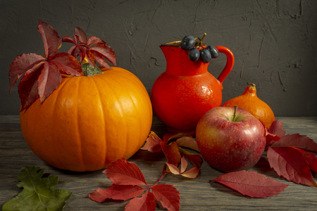 Two pumpkins with bright autumn leaves apple, blue grapes acorn and red jug on wooden rustic table against a black rough concrete grunge wall. Autumn Harvest and Holiday still life with free copy spaceの写真素材