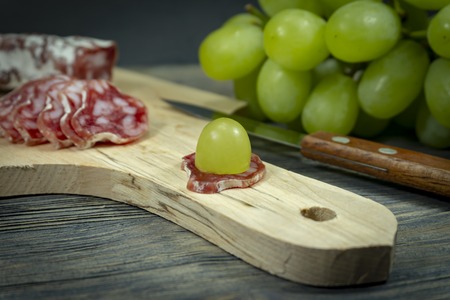 Sliced fresh green grape on salami on the handle of a wooden cutting board with sharp knifeの写真素材