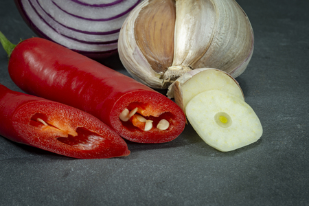 Close up one fresh red hot chili pepper onion and garlic cut to slices on background of black slate board, low angle side viewの写真素材