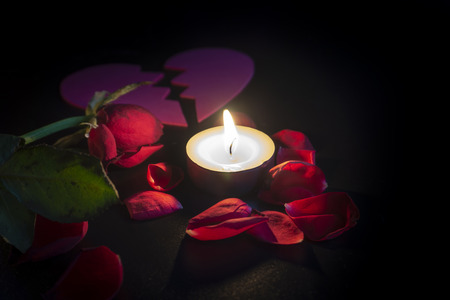 Tea candle and broken heart, red rose petals with shallow depth of focus with reflection on black backgroundの写真素材