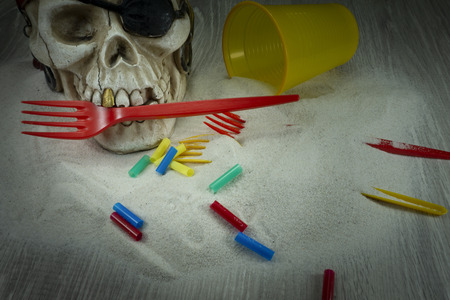 Pirate skull toy with plastic forks, straws, disposable glass and sand, viewed from high angle in close-up. Funny Jolly Roger, plastic waste problem conceptの写真素材