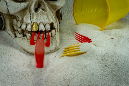 Pirate skull toy with red plastic fork in teeth, straws, disposable glass and sand, viewed from high angle in close-up. Funny Jolly Roger, plastic waste problem conceptの写真素材