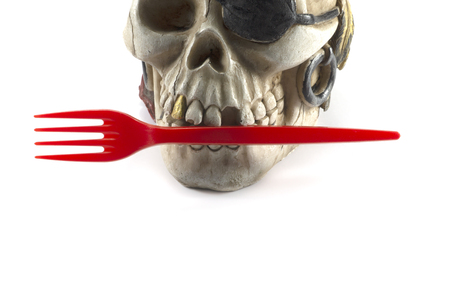 Pirate skull toy with red plastic fork in teeth, viewed in close-up against white background. Plastic waste problem conceptの写真素材