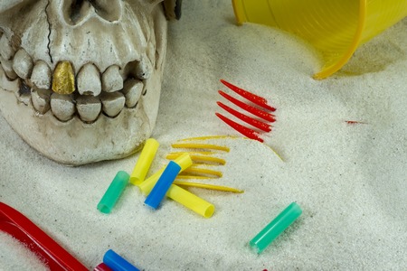 Pirate skull toy with plastic forks, straws, disposable glass and sand, viewed from high angle in close-up. Funny Jolly Roger, plastic waste problem conceptの写真素材