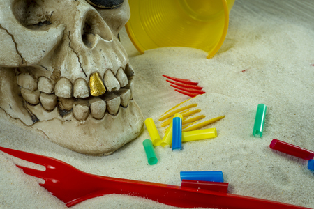 Pirate skull toy with plastic forks, straws, disposable glass and sand, viewed from high angle in close-up. Funny Jolly Roger, plastic waste problem conceptの写真素材