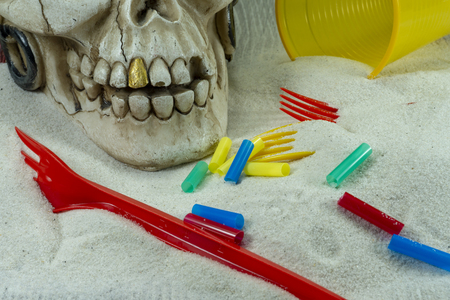 Pirate skull toy with plastic forks, straws, disposable glass and sand, viewed from high angle in close-up. Funny Jolly Roger, plastic waste problem conceptの写真素材