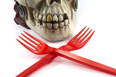 Pirate skull toy with red plastic fork in teeth, viewed in close-up against white background. Funny Jolly Roger or plastic waste problem conceptの写真素材