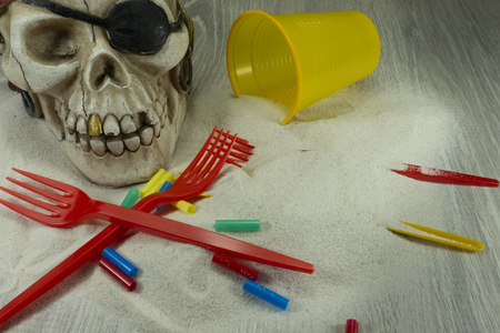 Pirate skull toy with plastic forks, straws, disposable glass and sand, viewed from high angle in close-up. Funny Jolly Roger, plastic waste problem conceptの写真素材