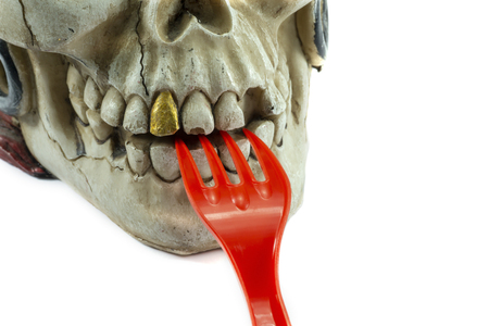 Pirate skull toy with red plastic fork in teeth, viewed in close-up against white background. Plastic waste problem conceptの写真素材