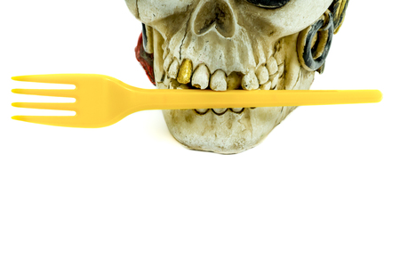 Pirate skull toy with yellow plastic fork in teeth, viewed in close-up against white background. Plastic waste problem conceptの写真素材