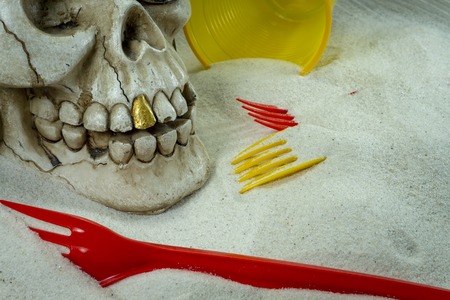 Pirate skull toy with plastic forks, disposable glass and sand, viewed from high angle in close-up. Funny Jolly Roger, plastic waste problem conceptの写真素材