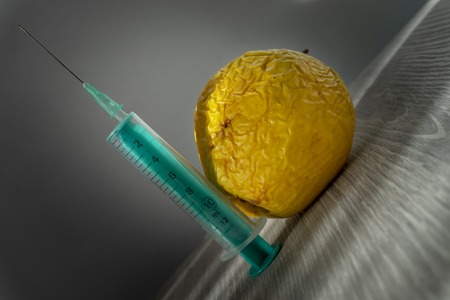 Old withered fresh apple with hypodermic needle inserted in a concept of healthcare, plastic surgery, treatment, research and medicine over grey backgroundの写真素材