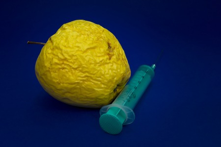 Old withered fresh apple with hypodermic needle inserted in a concept of healthcare, plastic surgery, treatment, research and medicine over a blue backgroundの写真素材