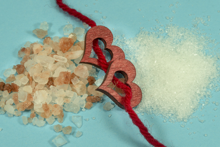 Two wooden decorative hearts threaded on red rope with a pile of rough salt crystals on one side and fine crushed salt or sugar grains on the other in a conceptual imageの写真素材