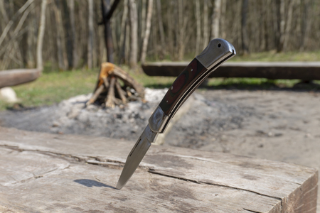 Folding knife stuck upright by the point in an old wooden picnic table at a campsite in a grassy clearing in a forest or woodland with a campfire built ready to light in the backgroundの写真素材
