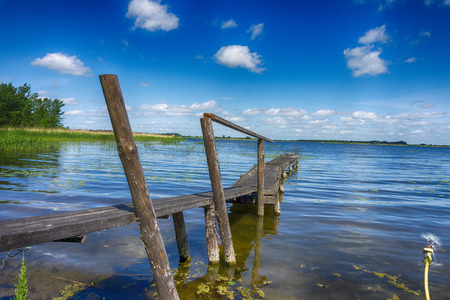 Old rickety wooden jetty leading into a calm lake reflecting the sunny blue sky with green shoreline and trees in the distanceの写真素材