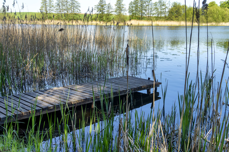 Old rustic wooden jetty on a tranquil lake with reeds and wild grasses on the bank and reflections on the waterの写真素材