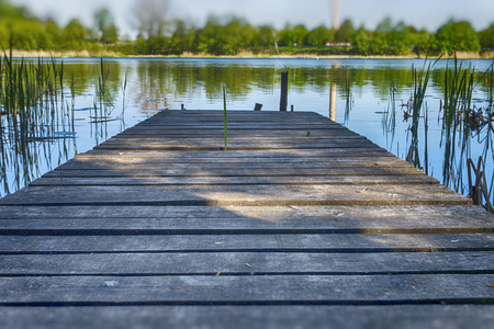 Old rustic wooden jetty on a tranquil lake with reeds and wild grasses on the bank and reflections on the waterの写真素材