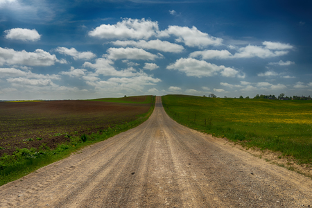 Gravel country road through farm fields and meadow in a spring landscape under a cloudy blue skyの写真素材