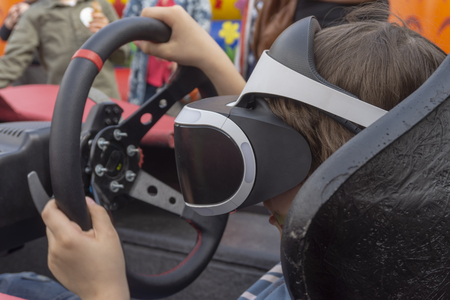 Child wearing VR glasses driving a red car in a playground turning the wheel as though cornering as he reacts with his virtual environmentの写真素材