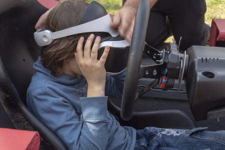 Child wearing VR glasses driving a red car in a playground turning the wheel as though cornering as he reacts with his virtual environmentの写真素材