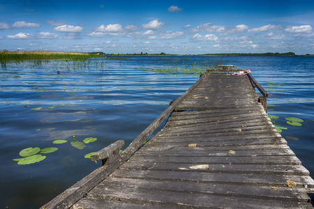 Old rickety wooden jetty leading into a calm lake reflecting the sunny blue sky with green shoreline and trees in the distanceの写真素材